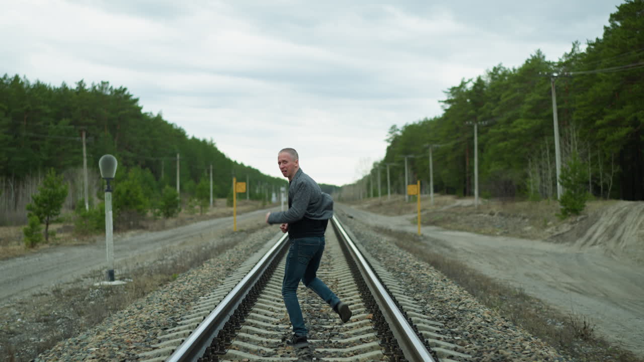 una vista cercana de un hombre anciano corriendo por una vía ferroviaria mientras mira hacia atrás, está vestido con un traje gris, vaqueros y zapatos de lona, con una vista de árboles y postes eléctricos