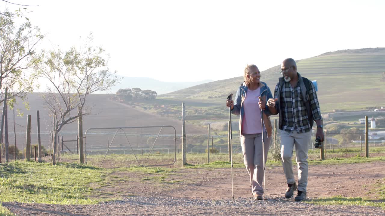 счастливая старшая афроамериканская пара с рюкзаками гуляет в солнечной природе, в замедленном движении