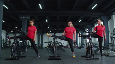 Gesunde-Kaukasische-Gruppe-Von-Frauen,-Die-Stretching-Auf-Einem-Stationären-Fahrrad-Im-Fitnessstudio-Ausüben