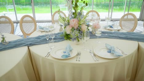 elegant wedding table setup with pastel floral centerpiece, blue napkins, and glassware, set in a bright indoor venue with a scenic green view