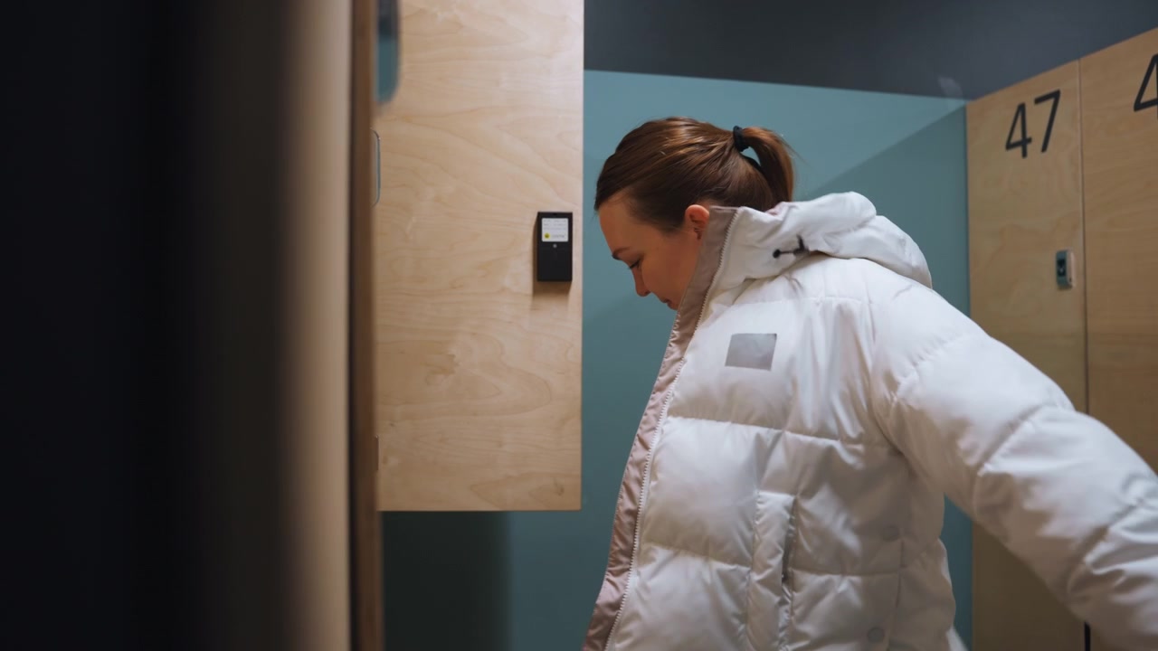 Locker room white woman removing coat after badminton session, wooden numbered lockers and teal wall, soft light and shadow, steady movements as she reaches inside locker, breathless relief and quiet