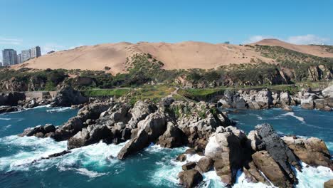 Traveling-Shot-from-Breaking-Waves-on-the-Rocks-of-the-Coast-with-Dunes-in-the-Background-and-some-buildings,-Viña-del-Mar,-Chile