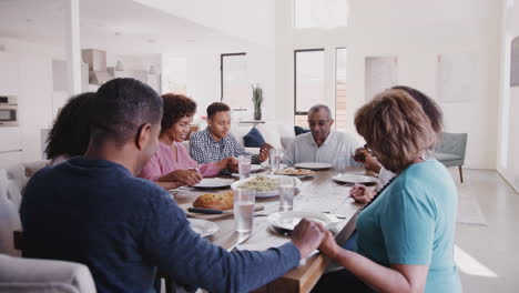 tres generaciones de una familia negra sentada en la mesa de cenar tomados de la mano y diciendo la gracia antes de la comida