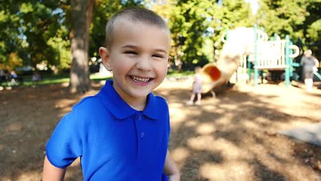 mischievous blonde boy in blue shirt laughing in the sunshine at the park