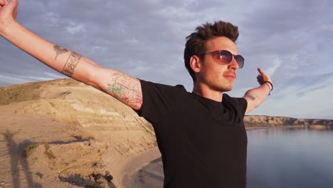 young man in black shirt wearing sunglasses standing with open arms on a mountain facing the sea at sunset - medium shot