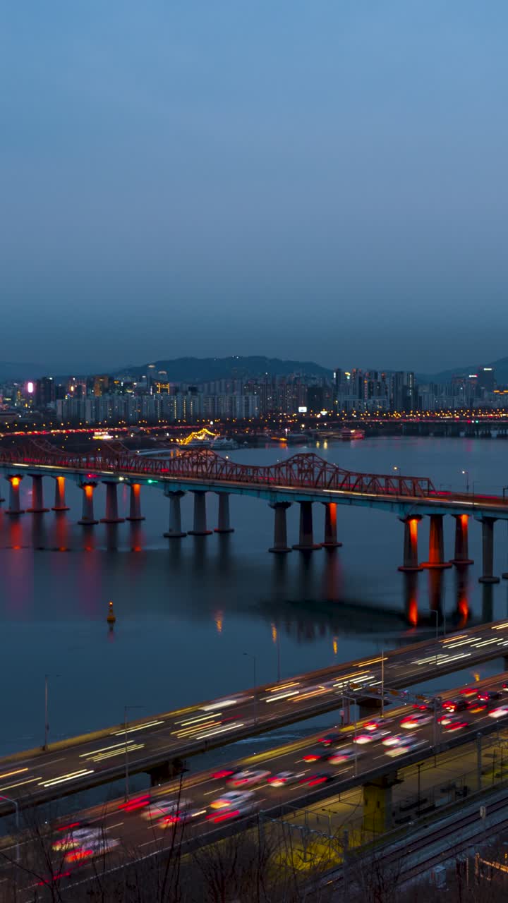 Timelapse verticale che riprende il passaggio dal giorno alla notte con scie luminose di traffico sull'autostrada Gangbyeonbuk-ro e l'iconico Ponte Dongho rosso riflesso nel tranquillo fiume Han.