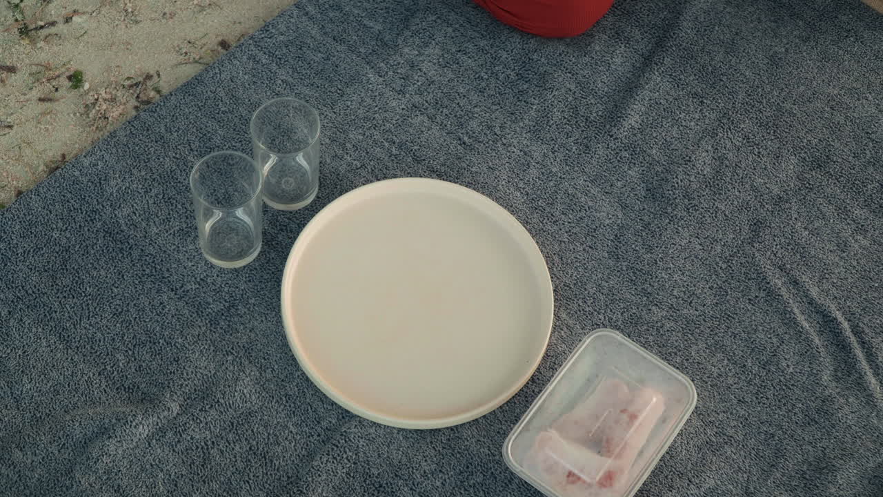 Manta de playa con preparación de té, pareja preparando un sereno picnic junto al mar con plato de cerámica, cuenco, vasos transparentes, aperitivos envasados y baguette, mano colocando una taza sobre una manta azul marino texturizada, luz solar suave.