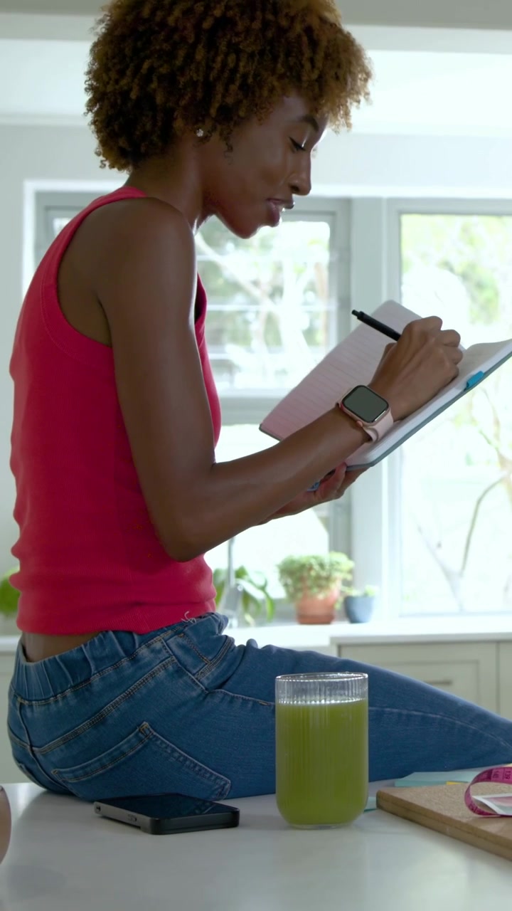 Vertikal video: Person kommer ind, kvinde i pink top og jeans gestikulerer, skriver i notesbog på køkkenbordet.