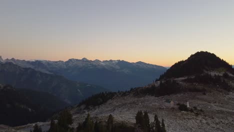 Beautiful-Aerial-View-at-Sunset-of-Lone-Cabin-on-Mount-Brew-Peak-with-Golden-Hour-Horizon-and-Pine-Trees-in-Canada-4K