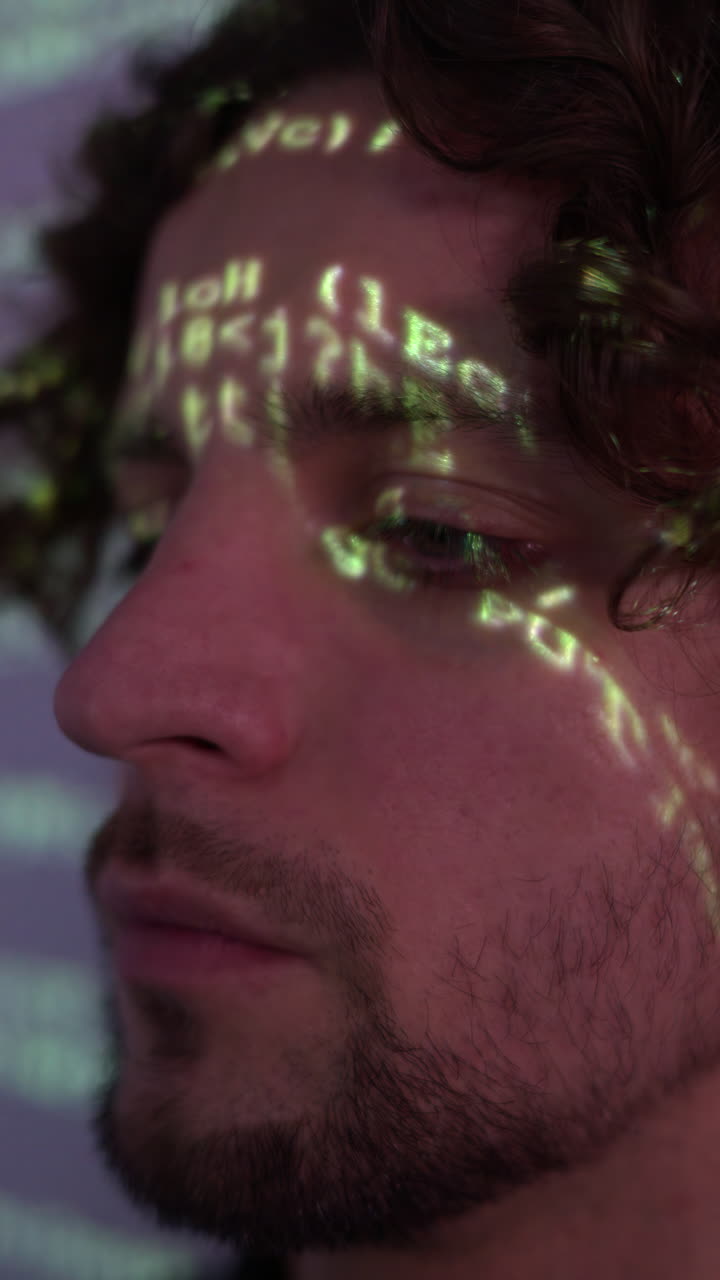 Man with curly hair in front of a projection of green computer code, illuminating on his face. Vertical