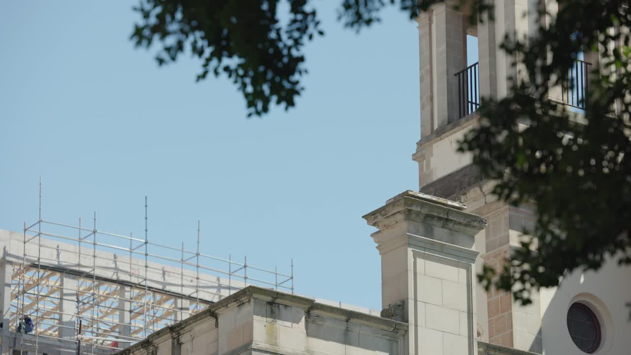 Brisa leve balança galhos enquanto trabalhador repara torre em andaimes durante renovação.