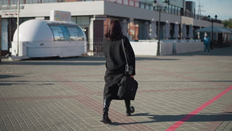 remote walker in black going to office, carrying briefcase and handbag, early morning light casting long shadows with a man walking close to the building, surrounded by modern architecture