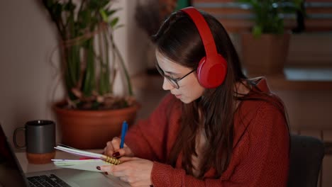 Confident-brunette-girl-in-wireless-headphones-and-glasses-takes-notes-in-her-notes-while-learning-foreign-languages-using-online-lessons-on-a-laptop-while-sitting-in-a-modern-kitchen-in-an-apartment