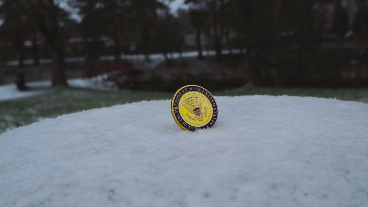 Footage that slowly pans and moves around a United States Presidential Seal coin that is standing up in white snow outdoors while snow falls on and around it during a winter cold day. Trees in back.