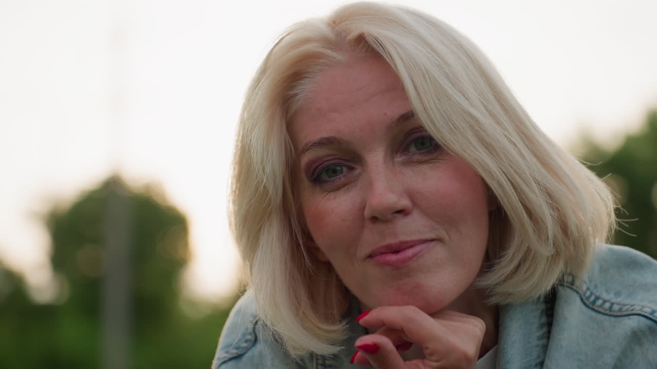 woman lying on blanket in park relaxing while adjusting hair with hand supporting head smiling gently blurred people playing nearby golden evening light casting soft glow over green grass