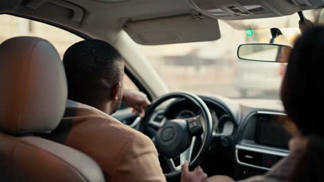 Vista-Trasera-De-Un-Hombre-De-Negocios-Feliz-Con-Piel-Negra-Con-Una-Chaqueta-Marrón-Viaja-En-El-Automóvil-Y-Conduce-El-Automóvil-Durante-Su-Viaje-De-Negocios-Con-Una-Colega-Chica-Morena-En-La-Ciudad.