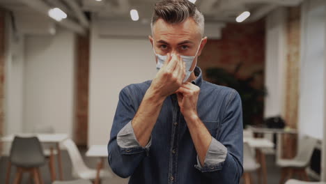 Mature-male-puts-on-a-medical-face-mask-to-work-inside-the-office.-Businessman-looking-at-camera,-front-view.