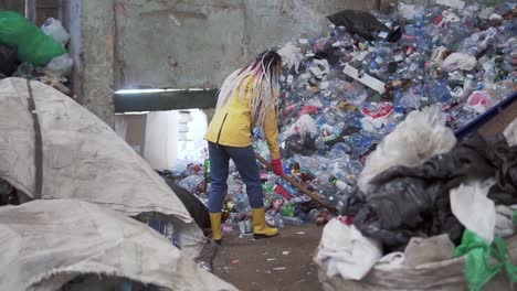 a rare view of a girl with dreadlocks, in yellow boots, scoops used bottles with a large duck shovel at a plastic recycling factory. huge pile of bottles on background