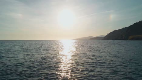 Bright-sun-over-the-sea,-aerial-drone-moving-backwards-to-the-rocky-beach