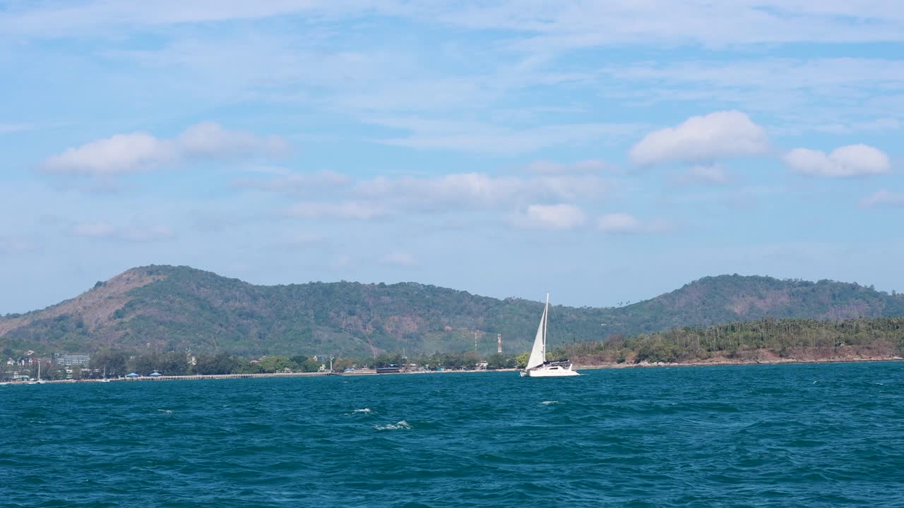 A sailboat glides through Phuket's ocean, framed by hilly jungle and clear skies, capturing serene maritime beauty
