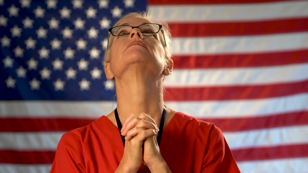 retrato medio ajustado de una enfermera de atención médica que parece preocupada y reza con una bandera estadounidense desenfocada