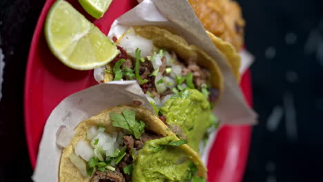 Traveling-close-up-shot-in-slow-motion-of-a-red-plate-with-three-different-tacos-served-with-slices-of-lime-and-guacamole-with-onion-and-cilantro-on-top