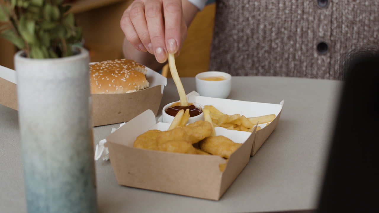 Person dipping french fry in ketchup with fast food meal