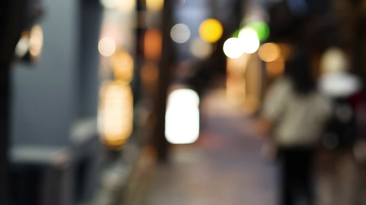 Out-of-focus figures walk through a softly lit, narrow urban passageway, creating a dreamy, atmospheric scene.