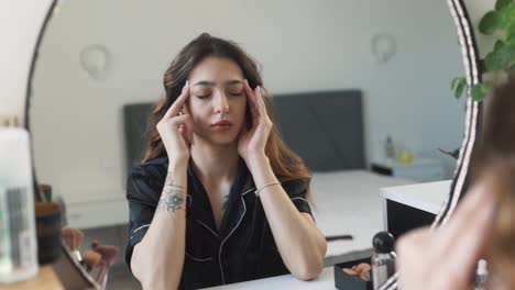 close-up portrait of a young beautiful woman in pajamas who woke up with a headache, massaging her temples in front of the mirror