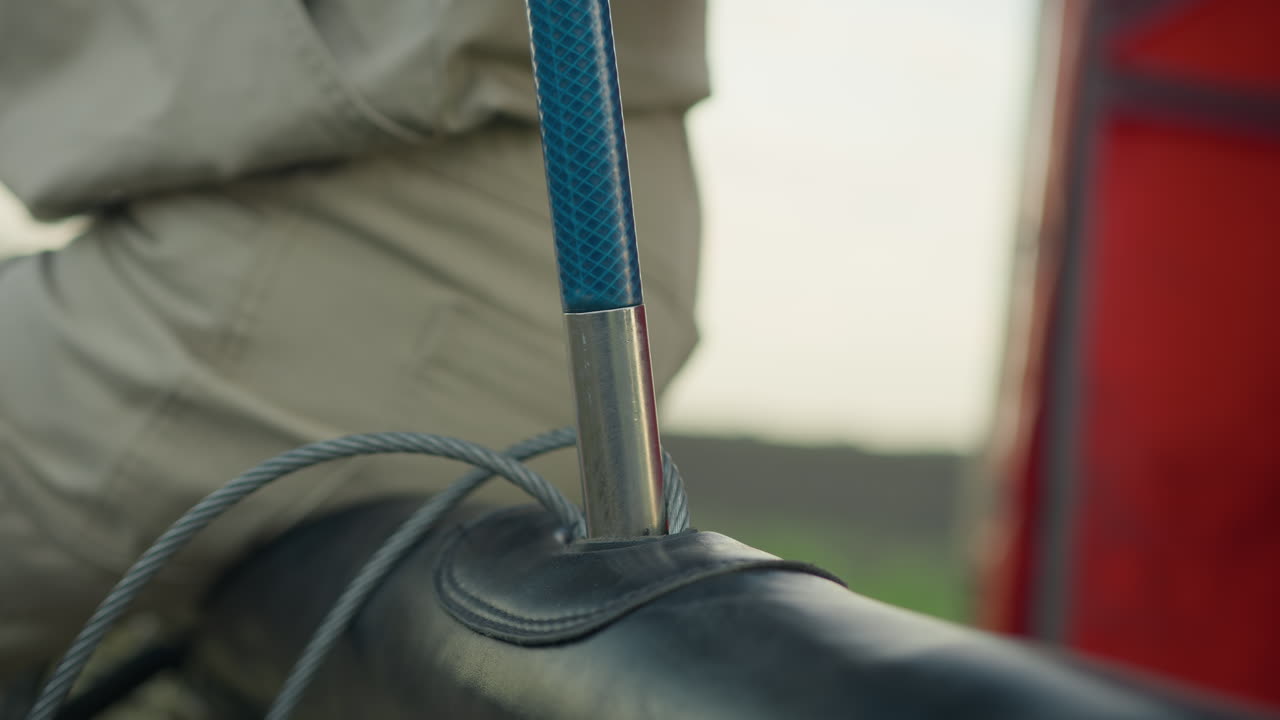 Gros plan sur une personne assise à côté d’un panier, alignant soigneusement une tige métallique verticale dans une douille ronde avec des câbles en acier enroulés reposant sur le bord, scène capturée à côté d’un camion rouge dans un champ ouvert