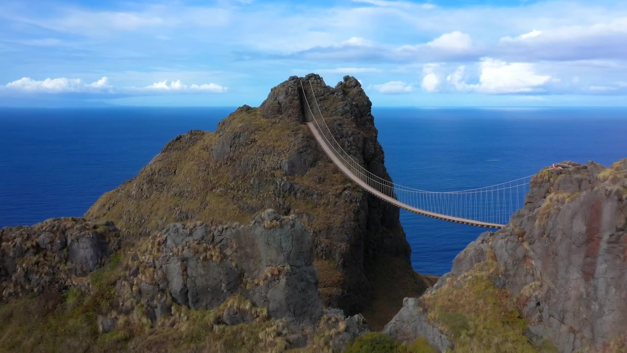 Passarela suspensa estreita com pranchas de madeira e corrimãos de cabo que se curva da beira do penhasco em direção a uma estreita abertura rochosa em um pico mais alto, pendurada bem acima do oceano azul profundo sob um céu suave parcialmente nublado