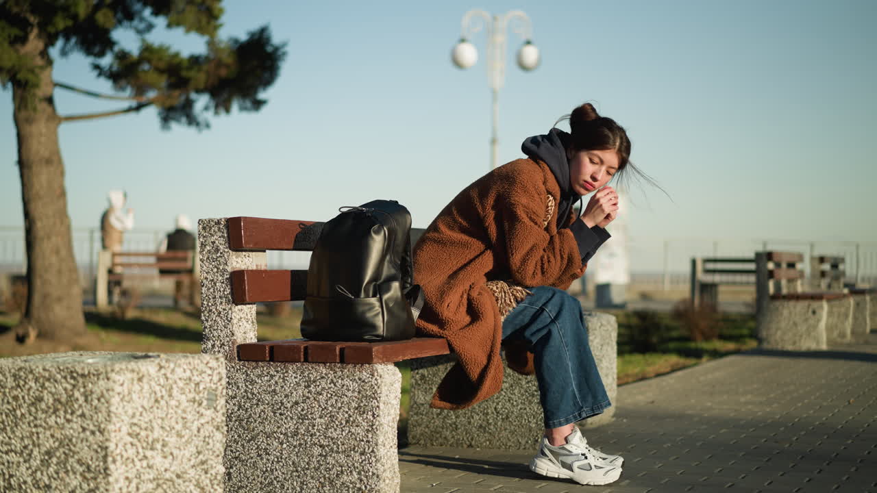 한 소녀가 도시 공원의 벤치에 혼자 앉아 있으며, 그녀의 머리는 그녀의 손에 머리를 고 깊이 우울해 보입니다. 그녀는 갈색 코트, 파란 청바지, 색 신발을 입고 있으며, 그녀의 에는 검은색 배이 있습니다.