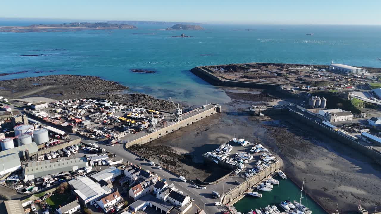 Guernsey St Sampsons harbour.Slow forward drone footage of harbour entrance and marina at low tide over industrial zone with views of Herm and Jethou in late afternoon sun with calm clear blue sea