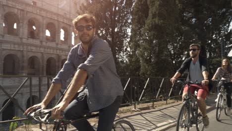 three young friends tourists riding bikes on road around colosseum in rome city centre on sunny day slow motion camera car steadycam