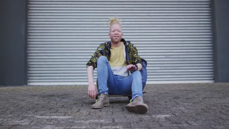 Portrait-of-smiling-albino-african-american-man-sitting-on-skateboard-looking-at-camera
