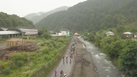 Amplia-Toma-Aérea-De-Un-Grupo-De-Ciclistas-De-Montaña-Cabalgando-Bajo-La-Lluvia-A-Través-Del-Barro-Junto-A-Un-Río.