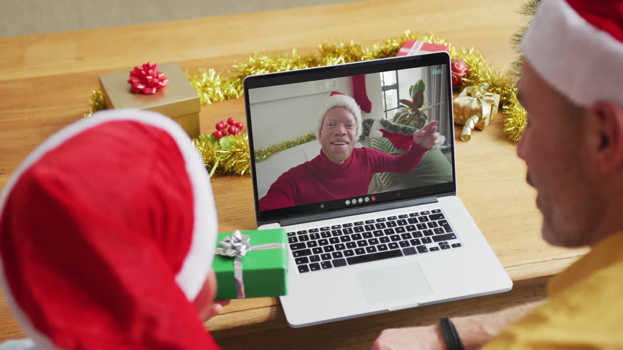 padre y hijo caucásicos con sombreros de santa usando una computadora portátil para una videollamada de navidad con un hombre en la pantalla