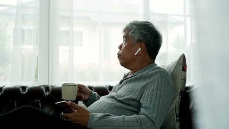 active senior man relaxing at home working, wearing bluetooth earbud enjoy listening to music from a mobile phone sitting comfortably on a sofa