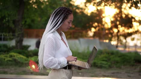 Lovely-woman-with-black-and-white-dreadlocks-hair-walking-by-the-street-with-open-silver-laptop,-typing.-Lens-flares-and-trees