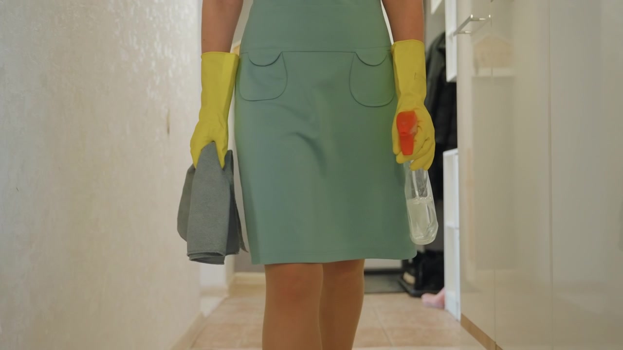 Woman entering hallway with cleaning cloth and spray bottle, yellow rubber gloves and green dress, brisk steps toward entrance, microfiber towel in hand, coat rack and shoes visible by doorway