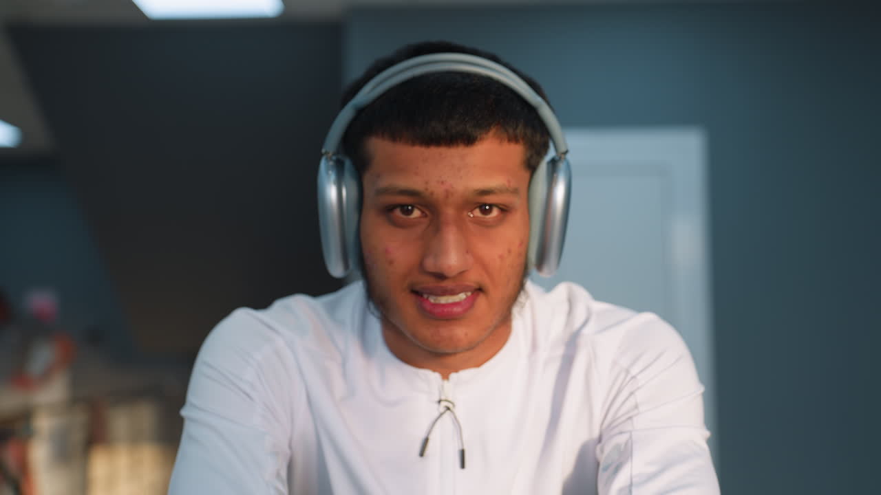 Cute man in white sports top and headphones cycles on stationary bike, smiling softly with door and blurred gym-goers in background under bright ceiling lights and modern interior