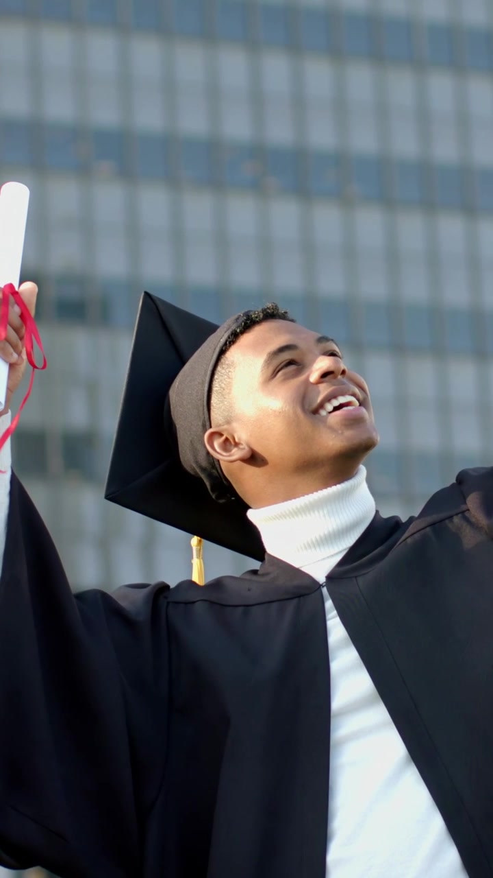 Video verticale: Telefono che entra nell'inquadratura, laureato sorridente in toga che solleva il diploma per una foto fuori dall'ufficio