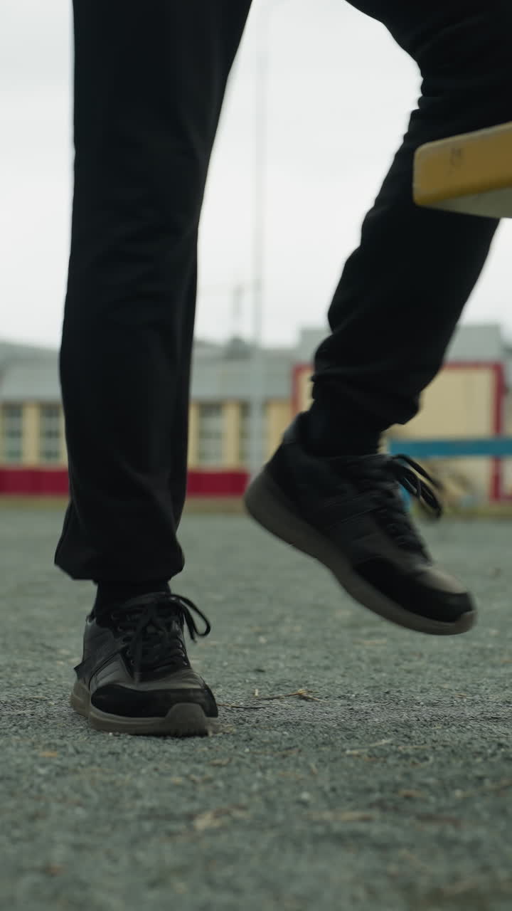 Close-up of a person's legs jogging slowly, wearing black jeans and black sneakers, near a bench with green grass underneath, a painted yellow house is visible in the distance