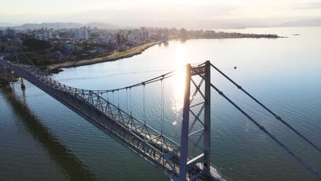 drone aerial scene of hercílio luz bridge at dusk at sunset with large capital city urban complex florianópolis late afternoon with metallic suspension bridge sun setting with bridge