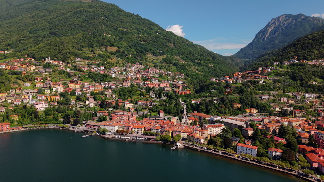 Drone pans from left to right along the Lake Como waterfront, showing red-roofed houses, trees, and the surrounding mountains under clear daylight