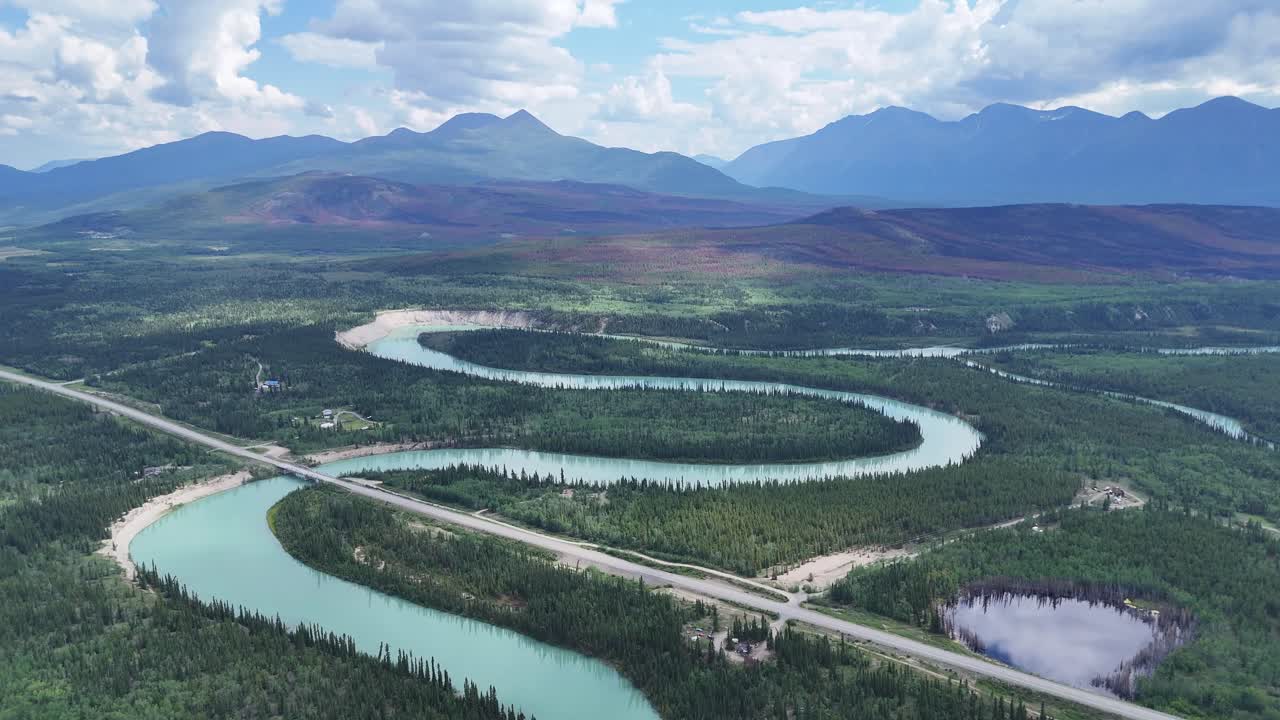 Stunning aerial view of a vibrant turquoise river winding through a lush green forest towards majestic blue mountains under a dynamic sky in Alaska