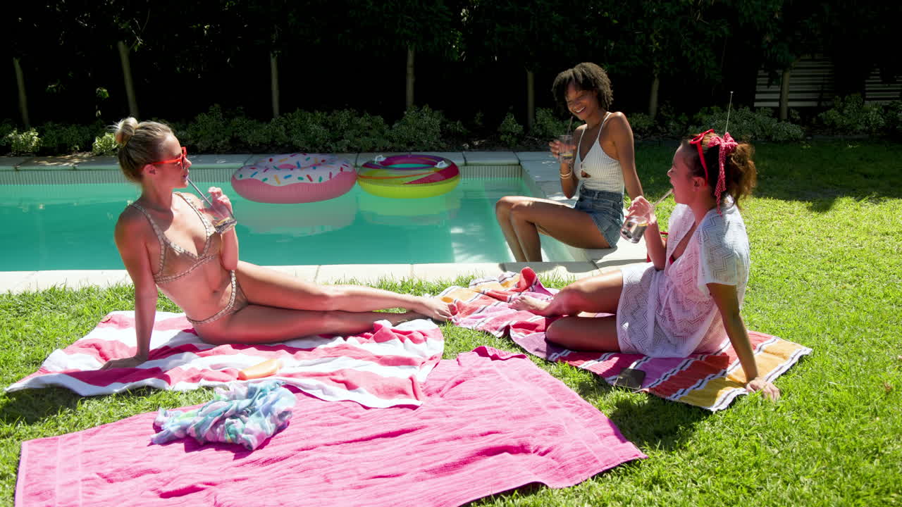 Diverse female friends relaxing by pool, enjoying drinks and laughter on sunny day