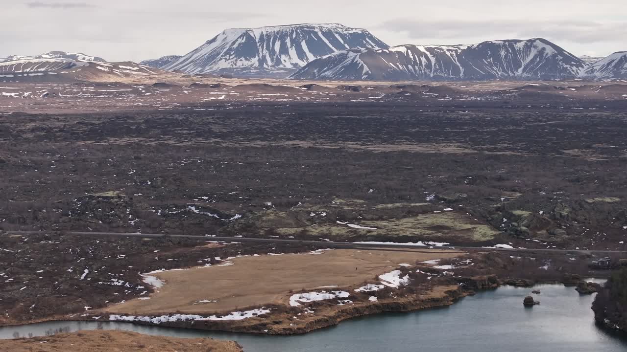 Panoramic aerial view of Lake Mývatn near Skútustaðir and Reykjahlíð, Iceland, with snow-capped volcanic mountains in the background and rugged lava fields surrounding the serene water body.