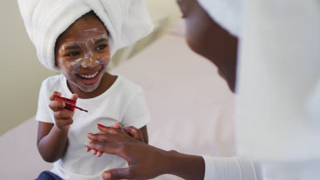 Happy-african-american-mother-and-daughter-sitting-on-bed-and-painting-nails