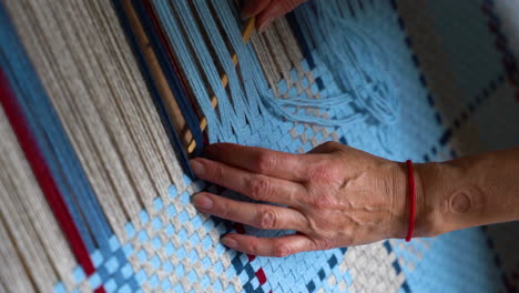 las manos de una anciana tejiendo hilos azules y beige en un telar